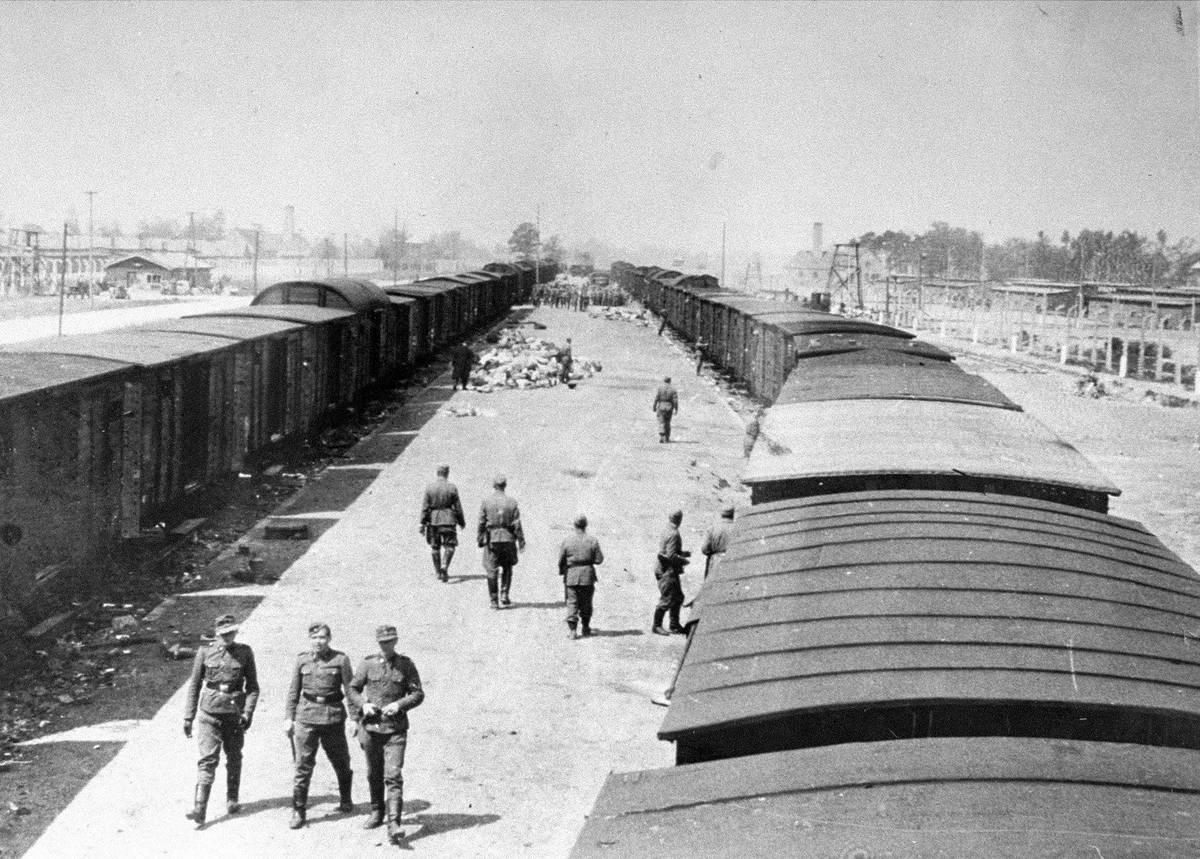 Květen 1944. Jeden z transportů je odbaven a čeká se na další "lidskou várku obětí". Stráže SS procházejí po příjezdové rampě tábora Auschwitz-Birkenau v Osvětimi. Rampa byla dokončena nedlouho před pořízením této fotografie. Deportační vlaky přijížděly dříve na rampu vzdálenou půl míle. Komíny krematorií II. a III. jsou vidět na horizontu.