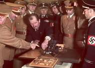 Dobový barevný snímek nejvyšších hodnostářů Třetí říše: jediným mužem bez uniformy je Ferdinand Porsche, předvádějící model jedné ze svých konstrukcí.